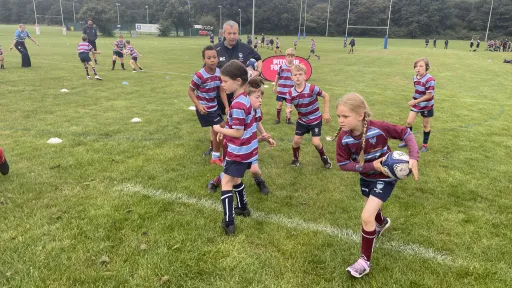 Young children in rugby uniforms practice on a grassy field with coaches nearby.