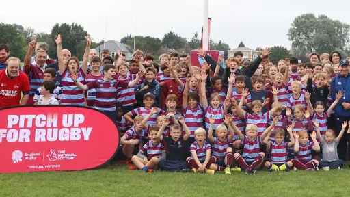 A large group of children and adults in rugby uniforms celebrating outdoors by a red banner.