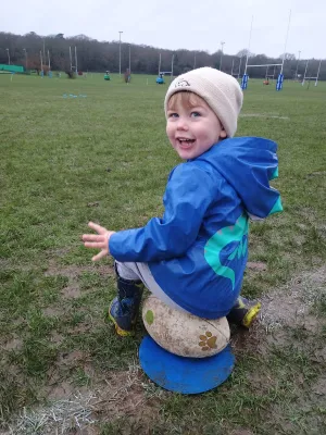 Child sitting on a rugby ball, smiling, wearing a blue jacket and beige beanie, on a grassy field at Rugger Rhinos.