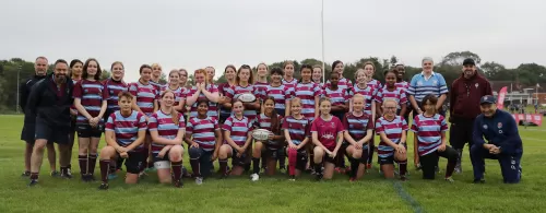 A group of rugby playing girls and coaches posing together on a grassy field, wearing striped uniforms.